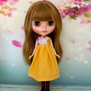 May include: A doll with long brown hair and a yellow dress. The doll has a white top with a colorful design and black boots. The background features pink and white floral elements.