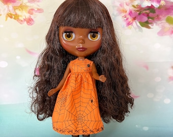 Puede incluir: Muñeca con cabello castaño y ojos grandes y amarillos. Viste un vestido naranja con un diseño de telaraña negra y botas negras. La muñeca tiene cabello largo y ondulado, sobre un fondo floral suave.