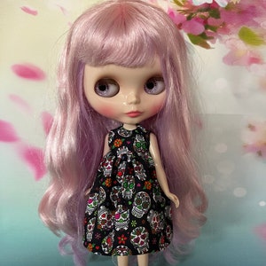 May include: A doll with lavender hair and large eyes wearing a black dress patterned with colorful sugar skulls. The doll is posed against a soft floral background, evoking a Day of the Dead theme.