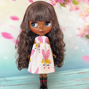 Può includere: Bambola con capelli castani e occhi blu, indossa un vestito bianco con stampa di coniglietti rosa e fiori. La bambola indossa anche stivali neri con dettagli rossi. Lo sfondo presenta un motivo floreale con colori rosa e bianco.