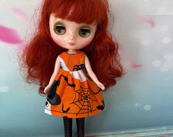 Puede incluir: Muñeca con pelo rojo y ojos verdes que lleva un vestido naranja con un estampado de telaraña y murciélagos negros. La muñeca también lleva botas negras con suelas rojas. Juguete con temática de Halloween.