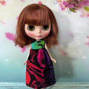 May include: A doll with auburn hair and green eyes wearing a colorful dress. The dress features a black background with a vibrant pattern of red, pink, and purple skeletal designs. The top of the dress is green, blue, and purple.