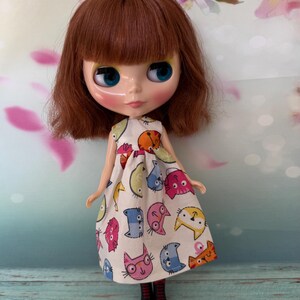 May include: A doll with auburn hair and blue eyes wearing a white dress patterned with colorful cartoon cat faces. The doll has a round face and is posed against a soft, floral background. The doll's dress has a sleeveless design.