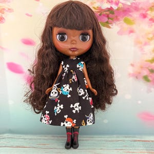 May include: A doll with brown skin and long, wavy brown hair. The doll wears a black dress with a white skull and crossbones pattern, black boots, and red and black striped socks. The background features pink and white floral elements.