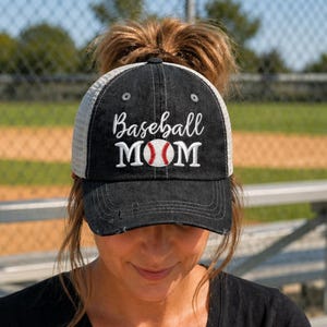 May include: Black baseball cap with a white mesh back and a distressed brim. The cap features the embroidered text "Baseball MOM" in white script, with a baseball graphic in the center. The cap is worn by a person.