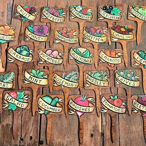 May include: A set of 24 wooden plant markers with hand-painted illustrations and names of various fruits, vegetables, and herbs. The markers are painted in bright colours and feature whimsical designs.