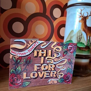 Könnte beinhalten: Eine Holzkarte mit einem roten, gelben und grünen Blumendesign. Die Karte trägt den Text "This is for lovers" in gelben Buchstaben. Die Karte ist signiert mit "Wolfman-Doherty."