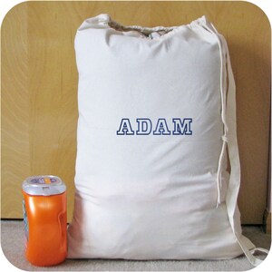 An off white drawstring laundry bag with the word 'ADAM' embroidered in blue Varsity NO Shadow font. A laundry detergent container is next to the bag.