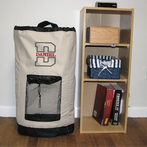 May include: A beige canvas laundry hamper with a black drawstring top and a black mesh pocket on the front. The hamper has a large gray letter "D" embroidered on the front with the name "DANIEL" in red letters below the "D". The hamper is next to a light wood shelf unit with three shelves. The top shelf has a wooden box, the middle shelf has a blue and white striped basket, and the bottom shelf has two books.