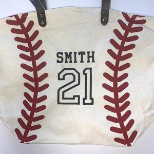 May include: A cream-colored canvas tote bag featuring a red baseball stitch pattern on both sides. The center of the bag displays the name SMITH and the number 21 in bold black lettering. The bag includes two dark brown leather-like handles.