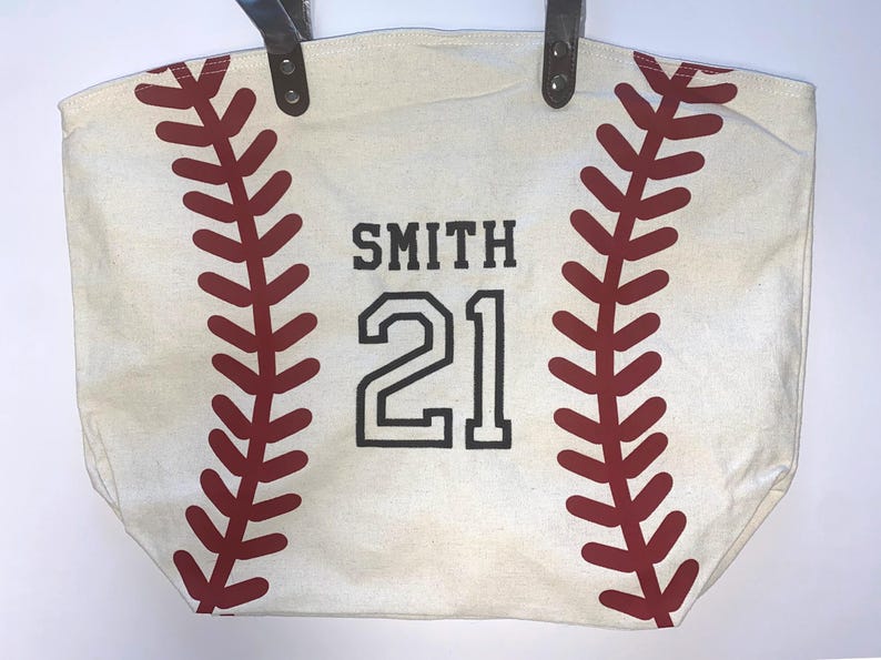 May include: A cream-colored canvas tote bag featuring a red baseball stitch pattern on both sides. The center of the bag displays the name SMITH and the number 21 in bold black lettering. The bag includes two dark brown leather-like handles.