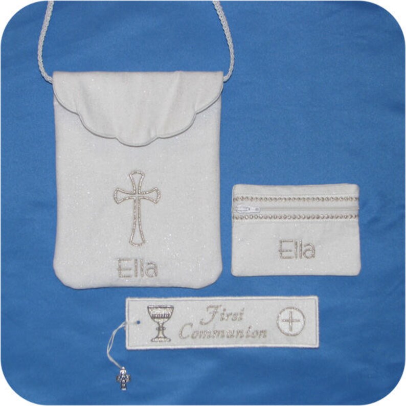 May include: A white First Communion set featuring a cross-adorned pouch with a scalloped flap, a small zippered coin purse, and a tag with the words "First Communion". The name "Ella" is embroidered on the pouch and purse.