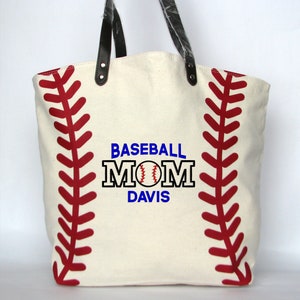 A large off white canvas tote bag with printed red baseball stitching on the sides. The bag has black faux leather handles attached with silver rivets. The front of the bag is personalized with a three line monogram:  top line is 'BASEBALL' in blue thread, middle line is 'MOM' in black thread and bottom line is  'DAVIS' in blue thread.  An embroidered baseball is replacing the 'O' in   'MOM' using the Block Mom Name design.