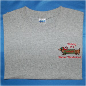 May include: Grey t-shirt with an embroidered dachshund wearing a Santa hat and sweater. The text "Walking in a Wiener Wonderland" is embroidered below the dachshund.