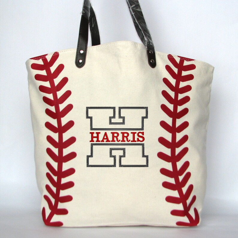An off white canvas tote bag with printed red baseball stitching on both sides. The bag has black faux leather handles and a large black embroidered letter 'H' with the word 'HARRIS' in red letters stitched in the middle of the 'H' using the Split Letter design.
