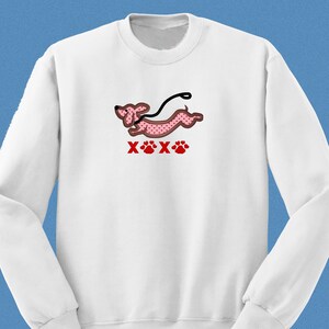 May include: White sweatshirt with a pink dachshund with red hearts printed on it. The dachshund is running with a leash and the text "XOX" is printed below the dog.