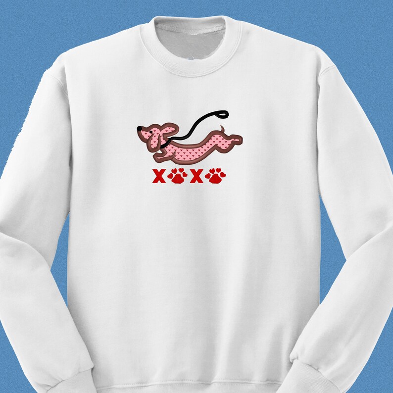 May include: White sweatshirt with a pink dachshund with red hearts printed on it. The dachshund is running with a leash and the text "XOX" is printed below the dog.