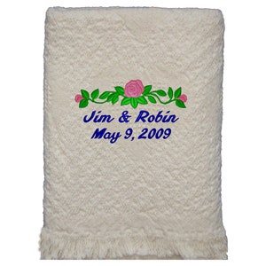 Natural colored cotton heart weave patterned throw blanket with fringe laying on wood background. Embroidery design on the blanket is 3 pink roses with leaves and couple's first names and wedding or anniversary date in blue thread using the Floral design.  The personalized design is located on the bottom center of the blanket and makes the perfect wedding or anniversary gift.
