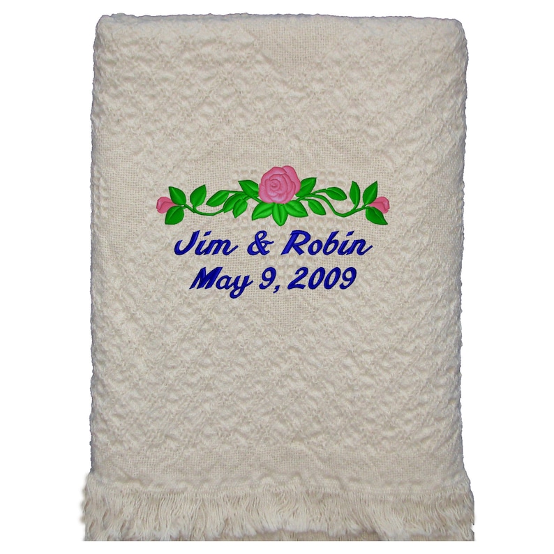 Natural colored cotton heart weave patterned throw blanket with fringe laying on wood background. Embroidery design on the blanket is 3 pink roses with leaves and couple's first names and wedding or anniversary date in blue thread using the Floral design.  The personalized design is located on the bottom center of the blanket and makes the perfect wedding or anniversary gift.