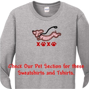 May include: A heather gray long-sleeve shirt featuring an embroidered dachshund with a pink heart pattern, a black leash, and the red text XOXO with paw prints. The shirt also includes the red text: Check Our Pet Section for these Sweatshirts and Tshirts.