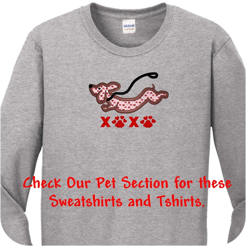 May include: A heather gray long-sleeve shirt featuring an embroidered dachshund with a pink heart pattern, a black leash, and the red text XOXO with paw prints. The shirt also includes the red text: Check Our Pet Section for these Sweatshirts and Tshirts.
