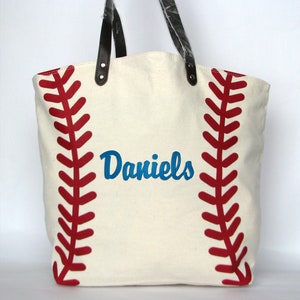 A large off white canvas tote bag with printed red baseball stitching on the sides. The bag has black faux leather handles and a blue embroidered name 'Daniels' in Brody script font.