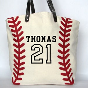 A large off white canvas tote bag with printed red baseball stitching on the sides. The bag has black faux leather handles and black embroidery that reads 'THOMAS' on the top line with a large '21' below using the  Athletic Number Name design.