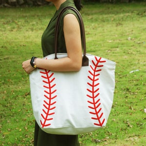 An off white canvas tote bag with printed red laces on both sides that resembles a baseball. The bag has faux leather handles which are hung over a baseball mom's shoulder to show how large the bag is.