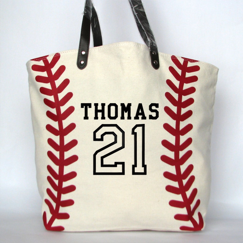 A large off white canvas tote bag with printed red baseball stitching on the sides. The bag has black faux leather handles and black embroidery that reads 'THOMAS' on the top line with a large '21' below using the  Athletic Number Name design.