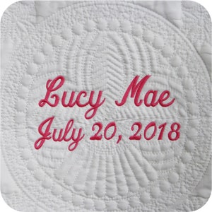 Pode incluir: Tecido acolchoado branco com texto bordado em rosa que diz "Lucy Mae July 20, 2018".
