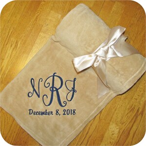 A soft tan fleece blanket with a satin ribbon tied around it. The ribbon has a hook & loop closure for easy gifting and/or quick storage. This wedding or anniversary throw blanket has large swirly script initials 'NRJ' and the date 'December 8, 2018' all in navy thread using the Marry Me design.  The personalization is located in the bottom corner of this fleece throw making an elegant and cozy wedding or anniversary gift.