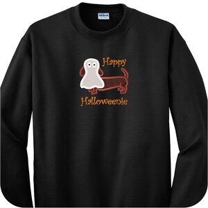 May include: A black long-sleeve sweatshirt featuring a graphic of a brown dachshund wearing a white ghost costume. The text "Happy Halloweenie" is printed in orange above and below the dog illustration. The garment has a crew neckline.