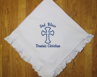May include: A white square blanket with a ruffled edge featuring blue embroidery. The center displays a blue cross with the text "God Bless" above it and the name "Dominic Christian" embroidered in blue script lettering below the cross.