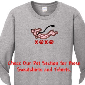 May include: A heather gray long-sleeve shirt featuring an embroidered dachshund with a pink heart pattern, a black leash, and the red text XOXO with paw prints. The shirt also includes the red text: Check Our Pet Section for these Sweatshirts and Tshirts.