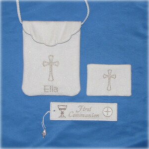 May include: A white First Communion set featuring a small bag with a cross and the name "Ella", a matching rectangular item with a cross, and a bookmark with "First Communion" text and a chalice illustration. The set is on a blue background.