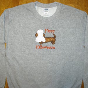 May include: A heather gray crewneck sweatshirt featuring an embroidered brown dachshund dog wearing a white ghost costume. Above the dog is the orange embroidered text Happy and below the dog is the orange embroidered text Halloweenie.