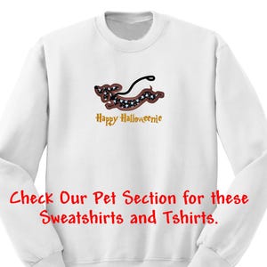 May include: A white long-sleeve sweatshirt featuring an embroidered dachshund dog with a black and white patterned body, a brown outline, and a black leash. Below the dog, the text reads Happy Halloweenie in gold. Red text at the bottom reads Check Our Pet Section for these Sweatshirts and Tshirts.