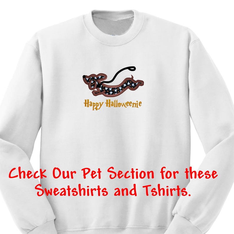 May include: A white long-sleeve sweatshirt featuring an embroidered dachshund dog with a black and white patterned body, a brown outline, and a black leash. Below the dog, the text reads Happy Halloweenie in gold. Red text at the bottom reads Check Our Pet Section for these Sweatshirts and Tshirts.
