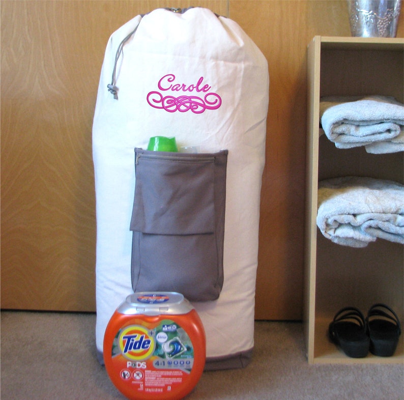 An extra, heavy duty large white laundry bag with gray trim and gray zippered pocket containing laundry detergent is in front of a closet. The laundry bag is monogrammed with Swirl Border design, consisting of a name "Carole" and a swirl below embroidered in fushia thread.