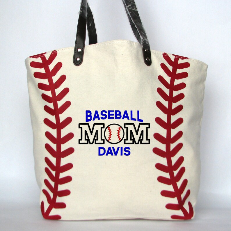 A large off white canvas tote bag with printed red baseball stitching on the sides. The bag has black faux leather handles attached with silver rivets. The front of the bag is personalized with a three line monogram:  top line is 'BASEBALL' in blue thread, middle line is 'MOM' in black thread and bottom line is  'DAVIS' in blue thread.  An embroidered baseball is replacing the 'O' in   'MOM' using the Block Mom Name design.