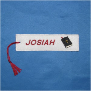 May include: White rectangular bookmark embroidered with the name "JOSIAH" in red letters. A small black book with a gold cross is on the right. A red tassel hangs from the top left corner. The background is blue.