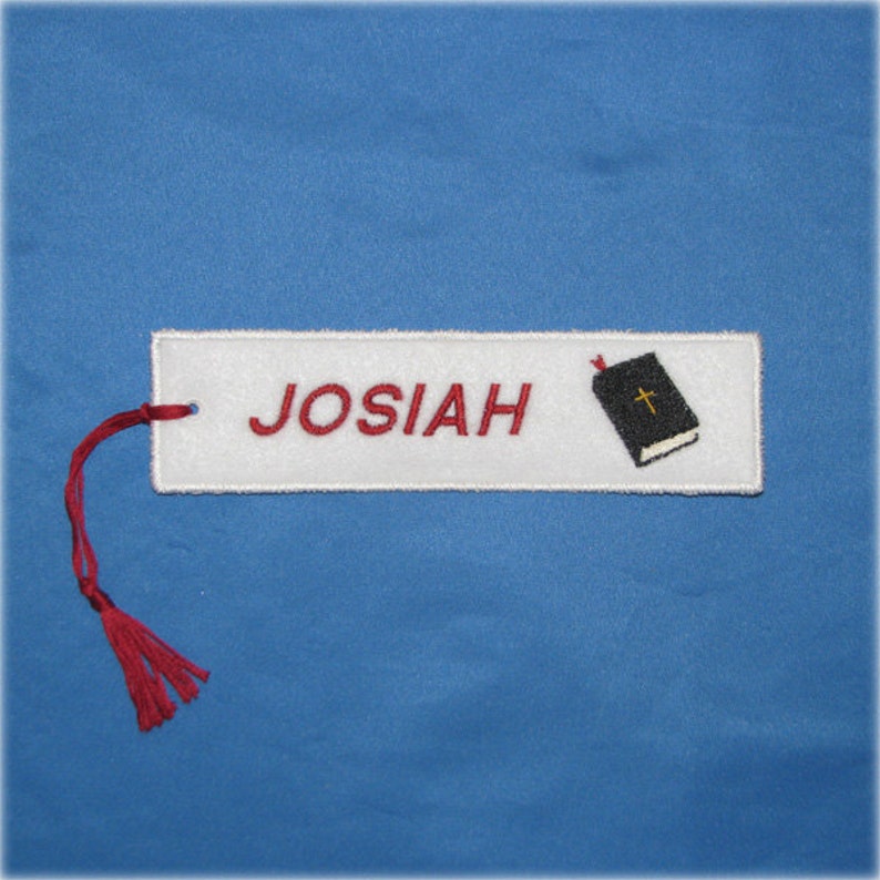 May include: White rectangular bookmark embroidered with the name "JOSIAH" in red letters. A small black book with a gold cross is on the right. A red tassel hangs from the top left corner. The background is blue.