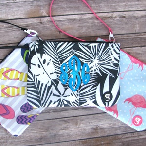 May include: Three rectangular zippered pouches with wrist straps. One features a black and white palm leaf print with a blue monogram, one has a striped pattern with colorful flip-flops, and one displays pink flamingos on a light blue background.
