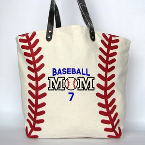 A large off white canvas tote bag with printed red baseball stitching on the sides. The bag has black faux leather handles attached with silver rivets. The front of the bag is personalized with a three line monogram:  top line is 'BASEBALL' in blue thread, middle line is 'MOM' in black thread and bottom line is  '7' in blue thread.  An embroidered baseball is replacing the 'O' in   'MOM' using the Block Mom Name design.