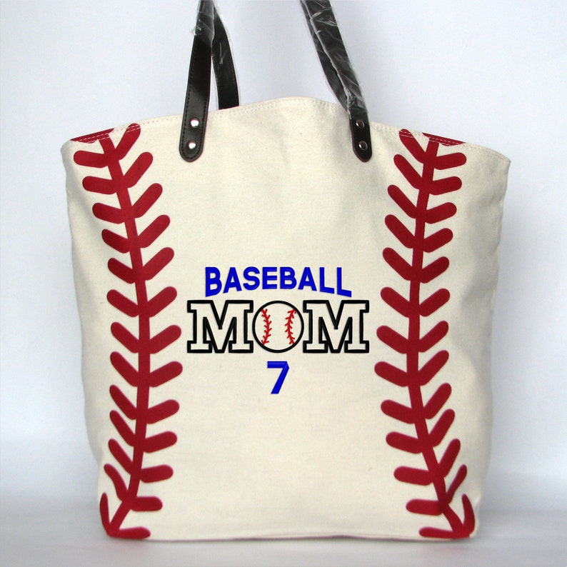 A large off white canvas tote bag with printed red baseball stitching on the sides. The bag has black faux leather handles attached with silver rivets. The front of the bag is personalized with a three line monogram:  top line is 'BASEBALL' in blue thread, middle line is 'MOM' in black thread and bottom line is  '7' in blue thread.  An embroidered baseball is replacing the 'O' in   'MOM' using the Block Mom Name design.