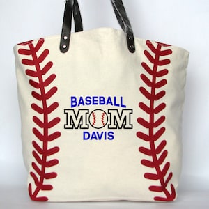A large off white canvas tote bag with printed red baseball stitching on the sides. The bag has black faux leather handles attached with silver rivets. The front of the bag is personalized with a three line monogram:  top line is 'BASEBALL' in blue thread, middle line is 'MOM' in black thread and bottom line is  'DAVIS' in blue thread.  An embroidered baseball is replacing the 'O' in   'MOM' using the Block Mom Name design.