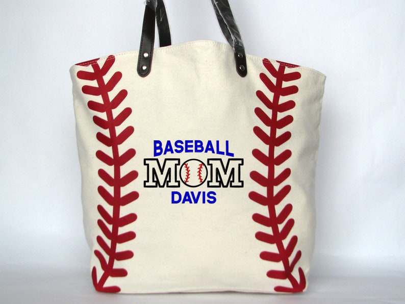 A large off white canvas tote bag with printed red baseball stitching on the sides. The bag has black faux leather handles attached with silver rivets. The front of the bag is personalized with a three line monogram:  top line is 'BASEBALL' in blue thread, middle line is 'MOM' in black thread and bottom line is  'DAVIS' in blue thread.  An embroidered baseball is replacing the 'O' in   'MOM' using the Block Mom Name design.