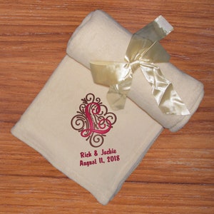 A soft vanilla fleece blanket with a satin ribbon tied around it. The ribbon has a hook & loop closure for easy gifting and/or quick storage. This anniversary or wedding throw blanket has large swirly red 'L' with brown swirl accents behind it and the text 'Rich & Jackie' and  'August 11, 2018' in red thread below using the Adorn Single design.  The personalization is located in the bottom corner of this fleece throw making an elegant and cozy wedding or anniversary gift.