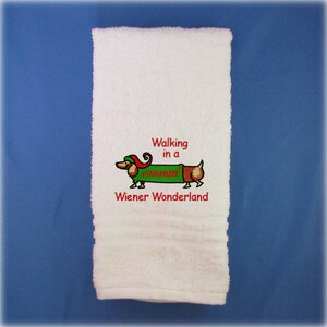 May include: White bath towel with red embroidered text that reads "Walking in a Wiener Wonderland". A cartoon dachshund wearing a green and red elf costume is embroidered above the text.