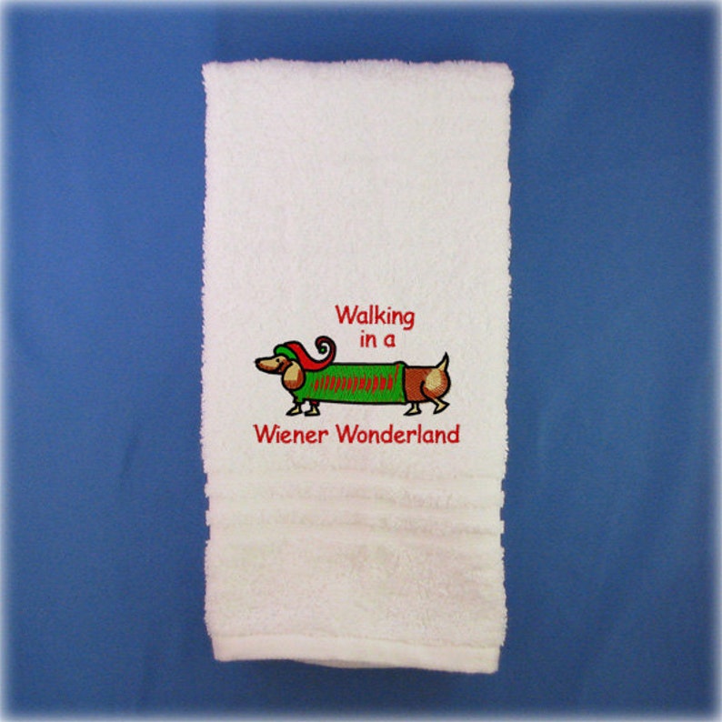 May include: White bath towel with red embroidered text that reads "Walking in a Wiener Wonderland". A cartoon dachshund wearing a green and red elf costume is embroidered above the text.